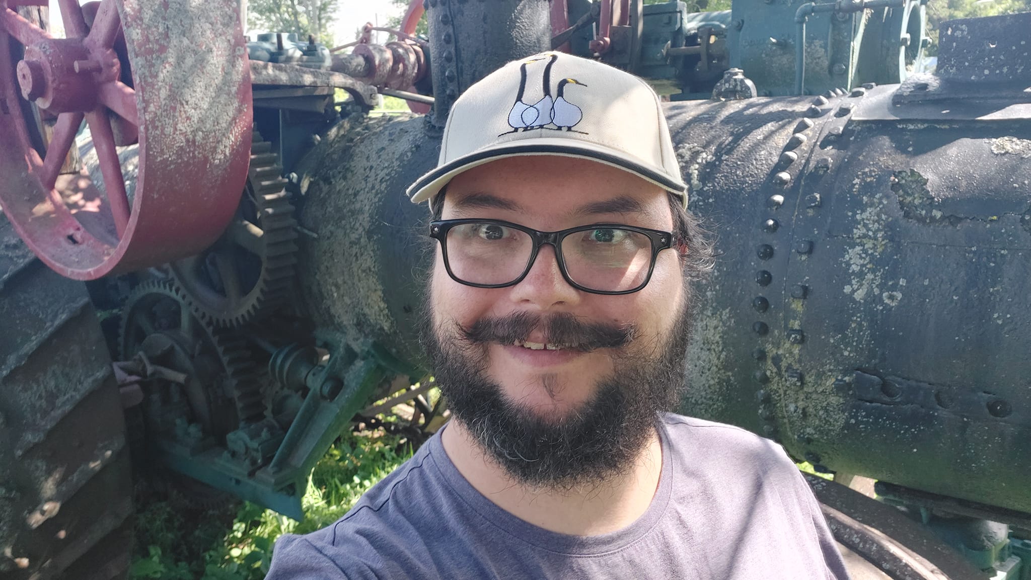 A man short hair and bushy facial hair excitedly stands in front of an old steam powered tractor